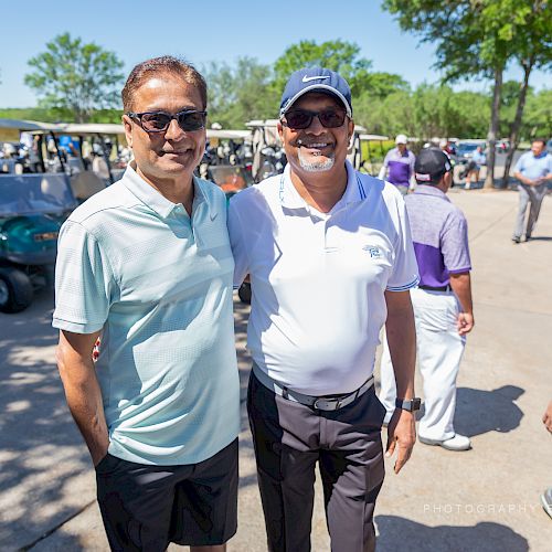 Two men are posing and smiling for a photo at a golf course, with golf carts and other people in the background.