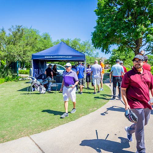 A golf event with people walking near tents on a sunny day, some holding golf equipment and others socializing.