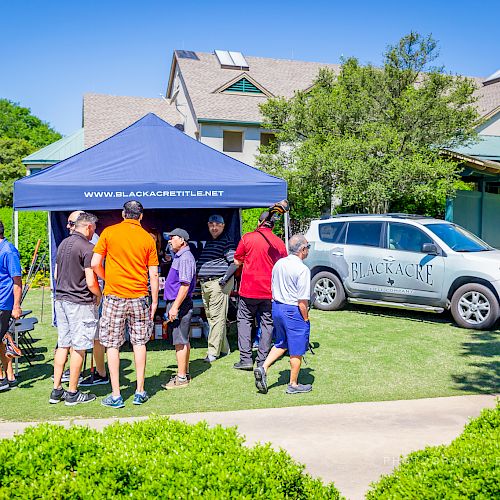 A group of people gather under a canopy tent labeled www.BlackacreTitle.com, next to a parked SUV and a building, on a sunny day.