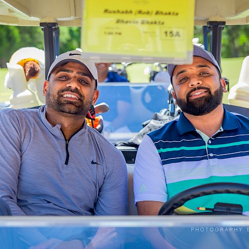 Two men are sitting in a golf cart, looking at the camera, with other people and golf carts in the background.