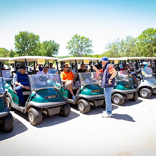 A group of people sits in golf carts lined up in a parking area, with a man standing and talking to them.