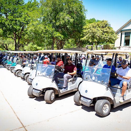 A group of people are sitting in golf carts lined up on a paved area, with trees and a building in the background.