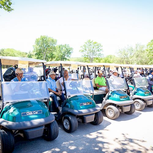 A group of people are sitting in parked golf carts, ready for an event on a sunny day at a golf course with trees in the background.