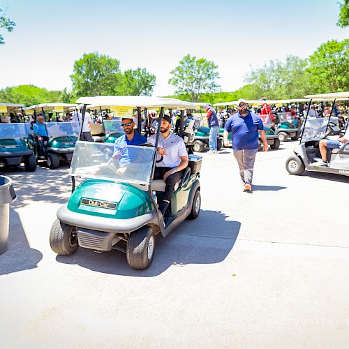 People riding and walking near a row of golf carts in a sunny outdoor area with green trees in the background, ending the sentence.