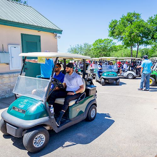 Two men in a golf cart in front of a building; others are in the background with more golf carts, and some greenery is visible.