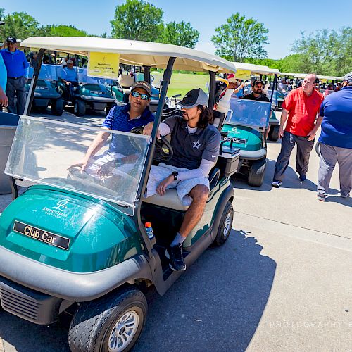 People are gathered outdoors, some in golf carts and others standing around, appearing to prepare for a golf event or activity.