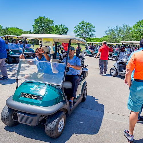 People are gathered around and riding golf carts in a sunny outdoor area. A person in an orange shirt is standing and chatting with others.