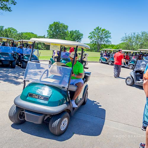 People are gathered around golf carts on a sunny day, preparing for a game of golf at what appears to be a golf course.