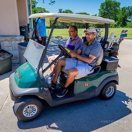 Two men riding a golf cart on a sunny day, with golf clubs in the back. They are at a golf course with green surroundings and clear skies.