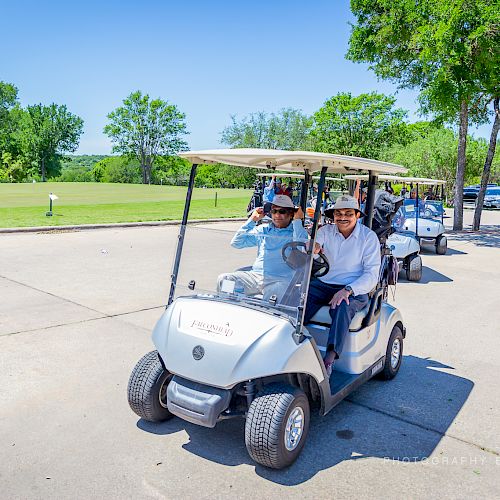 People riding in and standing near golf carts on a sunny day, with greenery and a blue sky in the background.