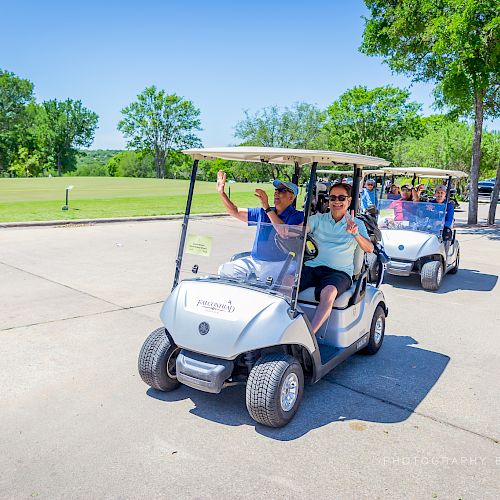 Two golf carts are being driven on a paved path with people in casual attire, surrounded by greenery and trees on a sunny day.