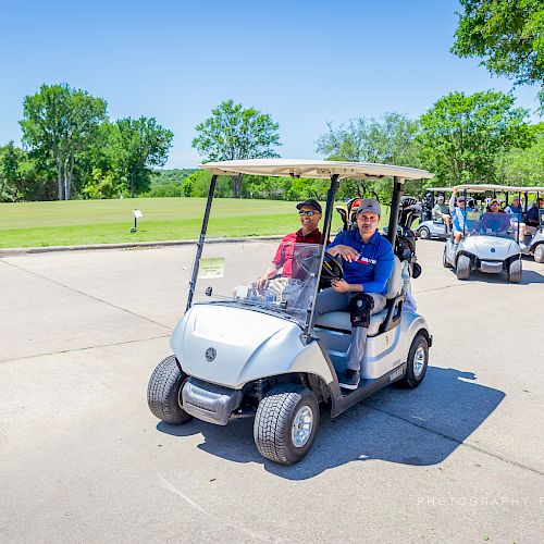 A group of people driving golf carts on a sunny day near a golf course with green trees and a clear blue sky in the background.