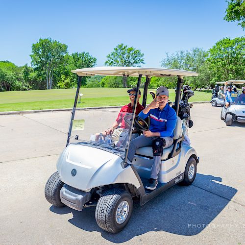 A group of people ride in golf carts on a sunny day at a golf course, with greenery and trees in the background.