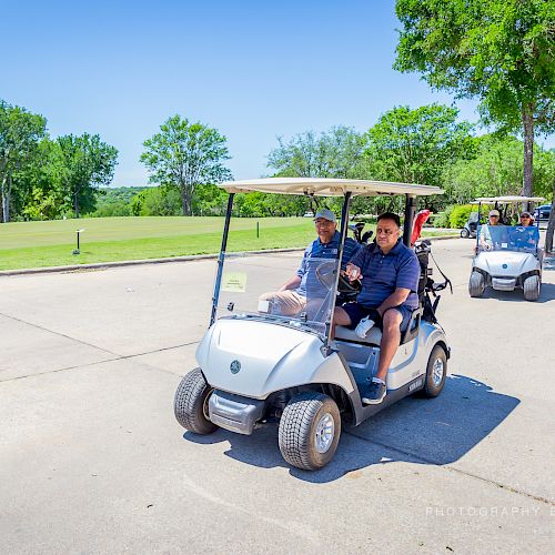 Two men are driving a golf cart on a paved path, with another golf cart following behind on a sunny day.