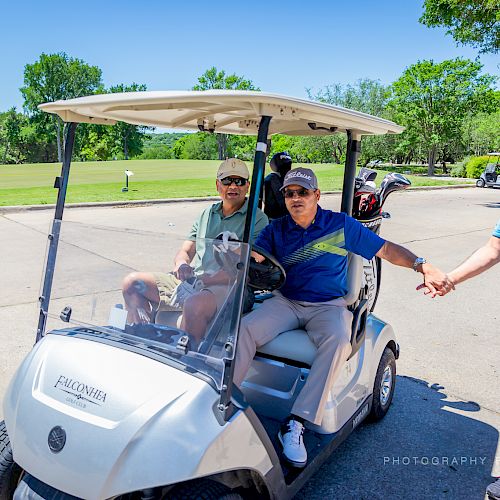 Three men, one in a golf cart and two standing outside. Two are shaking hands near a golf course on a sunny day.