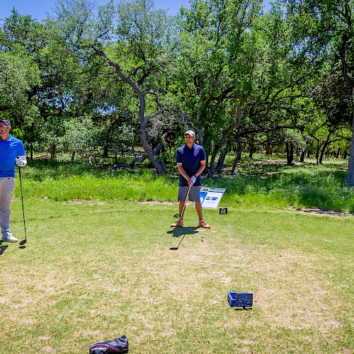 Two people are on a golf course, one preparing to swing while the other waits. Trees surround the area under a clear sky.