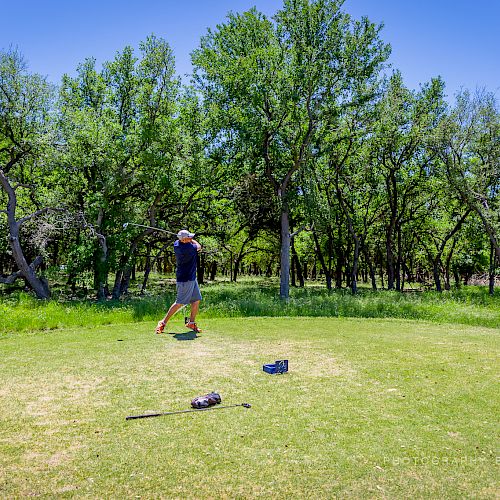 A golfer is mid-swing on a lush green course with trees in the background. Golf equipment lies on the grass.