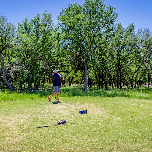 A person in a hat and shorts stands on a golf course, surrounded by trees. Golf clubs lie on the grass in the foreground, under a sunny sky.