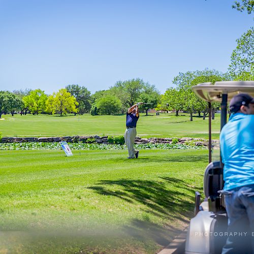 A golfer is swinging on a lush green course while two people ride in a golf cart in the foreground, surrounded by trees and under a clear sky.