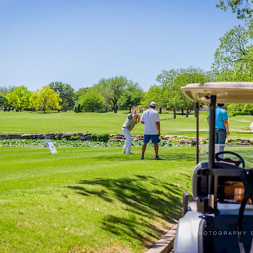 A group of people playing golf on a sunny day with a golf cart in the foreground and a scenic green landscape in the background.