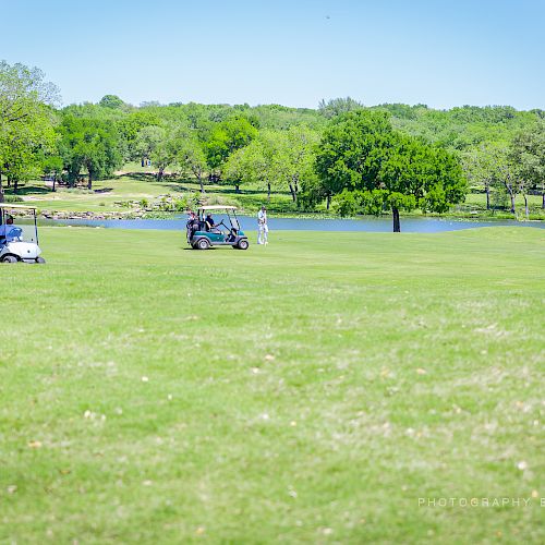 A green golf course with two golf carts, a few people in the distance, and a scenic lake surrounded by trees in the background.