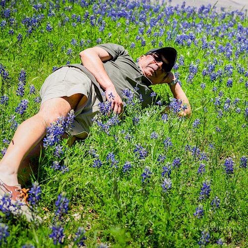 A person is lying in a field of blooming purple flowers, wearing casual clothes, a cap, and sunglasses. The background is filled with more flowers.