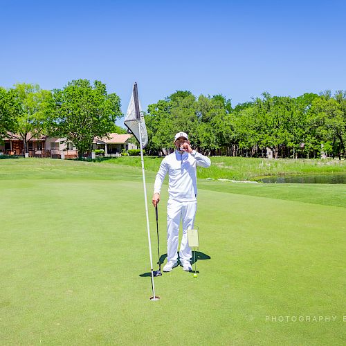 A person in white clothing stands on a golf course, holding a flag and talking on the phone, with houses and trees in the background.