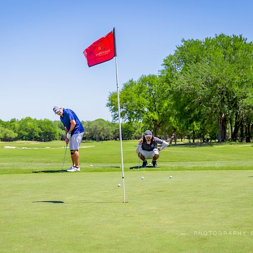 Three people on a golf course with one person putting near the hole while the others observe. The red flag marks the hole.