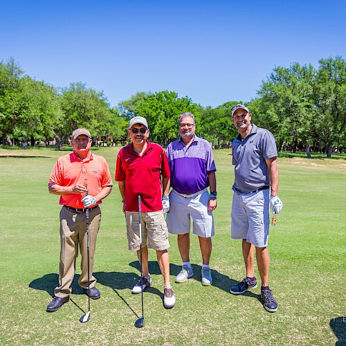 Four men in golf attire are standing on a golf course holding clubs, with trees and a golf cart in the background.