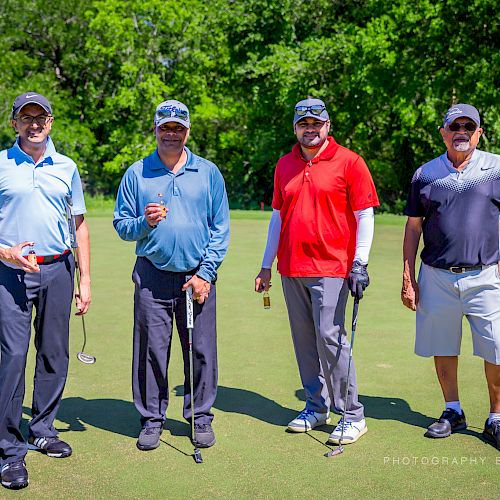 Four men are posing on a golf course, each holding a golf club, surrounded by greenery.