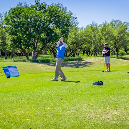 A golfer is swinging a club on a course while three people watch in the background. Trees and blue sky are visible.