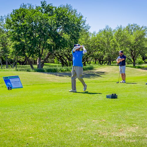 A golfer in a blue shirt is taking a swing on a green golf course while a group of people watches in the background.