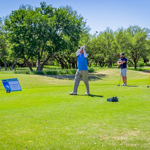 A golfer in a blue shirt takes a swing while others watch on a sunny golf course, with a sign and a box on the green.