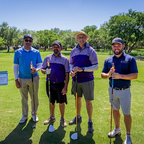 Four men are standing on a golf course, holding golf clubs. They are posing for a photo, dressed in casual golf attire, and the weather is sunny.