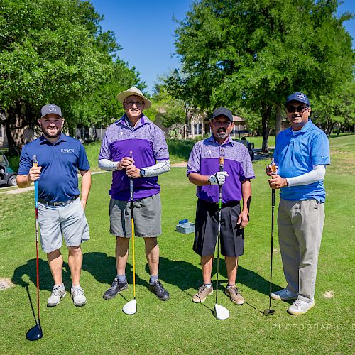 Four people standing on a golf course, holding golf clubs, posing for a photo on a sunny day.