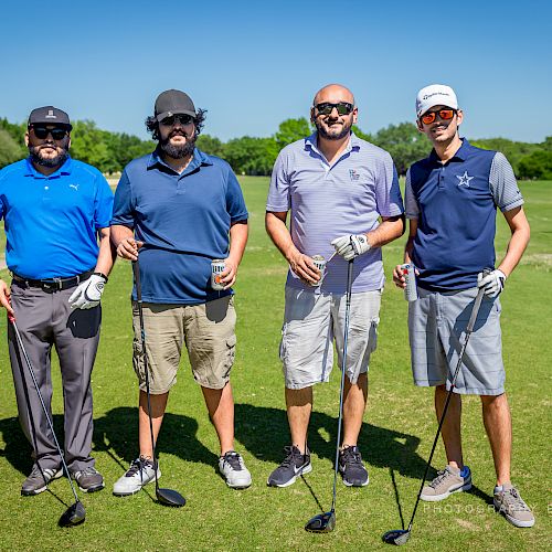 Four men standing on a golf course, holding golf clubs, wearing casual golf attire, and posing for the photo under a clear blue sky.