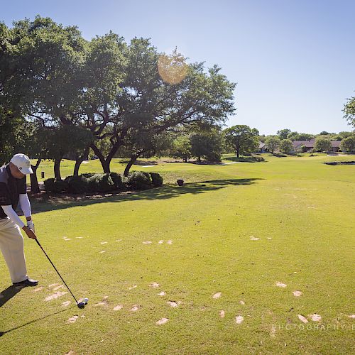 A person is preparing to hit a golf ball on a sunny golf course surrounded by trees and a manicured lawn.