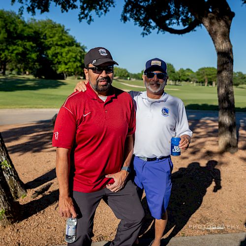 Two men are standing outside on a golf course, smiling, with trees and green grass in the background.