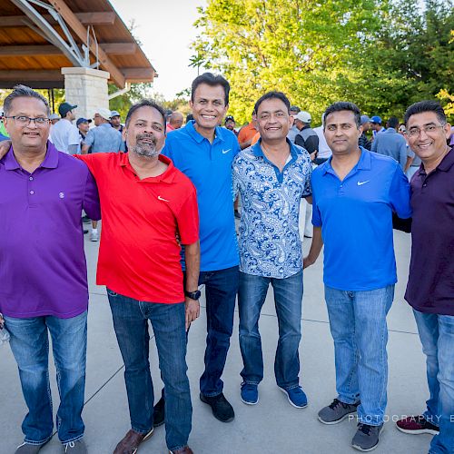 A group of six men are standing outdoors, smiling for the photo. The background features trees and a structure, with others visible in the distance.