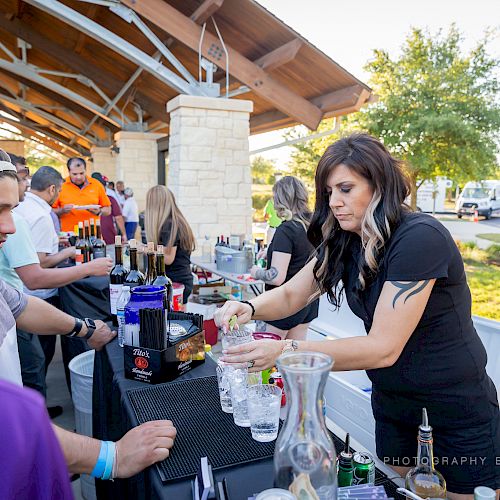 A group of people is gathered outdoors at a bar counter or event with a woman in black serving drinks, and various bar supplies are on the counter.