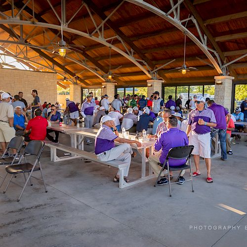 People gathered in an outdoor pavilion, sitting and standing around tables, socializing, and eating, in a spacious, well-lit setting.