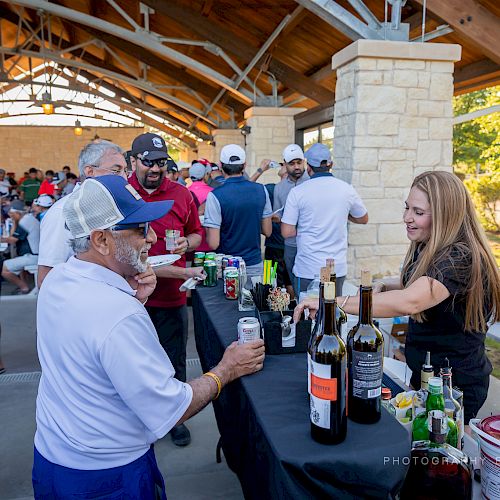 People are gathered at an outdoor event with drinks being served by a bartender at a covered pavilion.