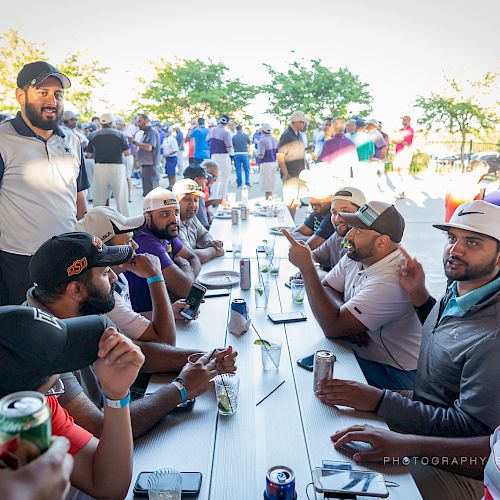 A group of people wearing hats and casual clothing are sitting at long tables outside, engaging in conversation and enjoying drinks.