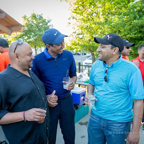 Several people are socializing outdoors, with two men in the foreground holding drinks and having a conversation. They seem to be at an event.