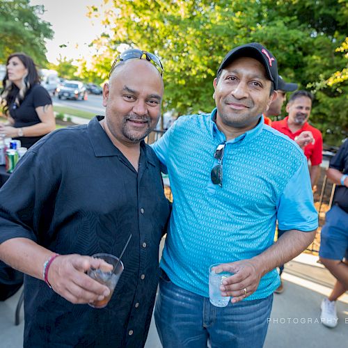 Two men are at an outdoor gathering with one holding a drink, posing for a photo while others socialize in the background.