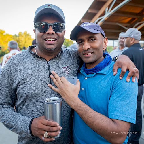 Two men smile, with one holding a drink in a gathering outdoors. The background shows others in casual attire. The image credits a photographer.