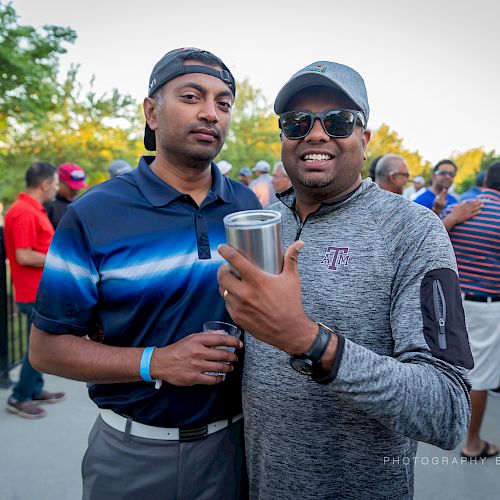 Two men standing together outdoors, smiling, both holding drinks. They are surrounded by a group of people socializing in the background.