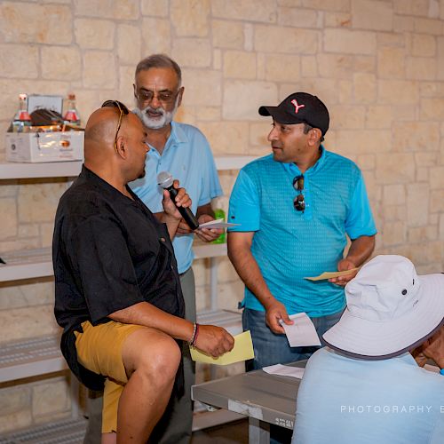 A group of individuals are engaged in a discussion indoors, with one holding a mic. A table with items and shelving is in the background.