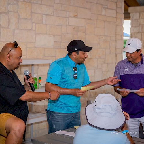 Three people are interacting in an outdoor setting; one is holding a microphone, and the others appear to be exchanging something.