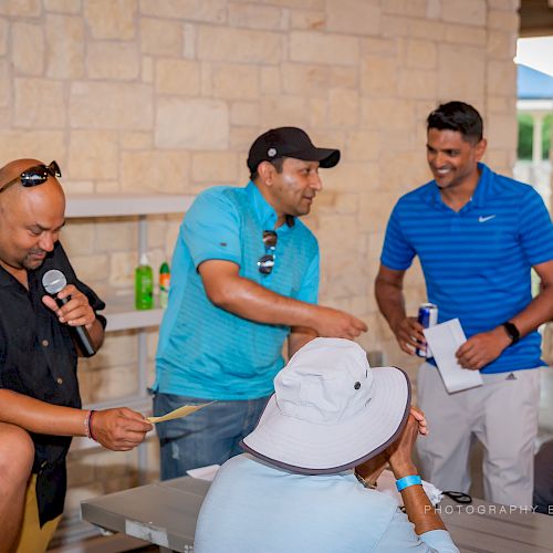 A group of people is interacting while one man speaks into a microphone. They seem to be having a friendly gathering.